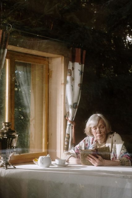 Seniorka číta pri otvorenom okne, na stole kanvica a šálka